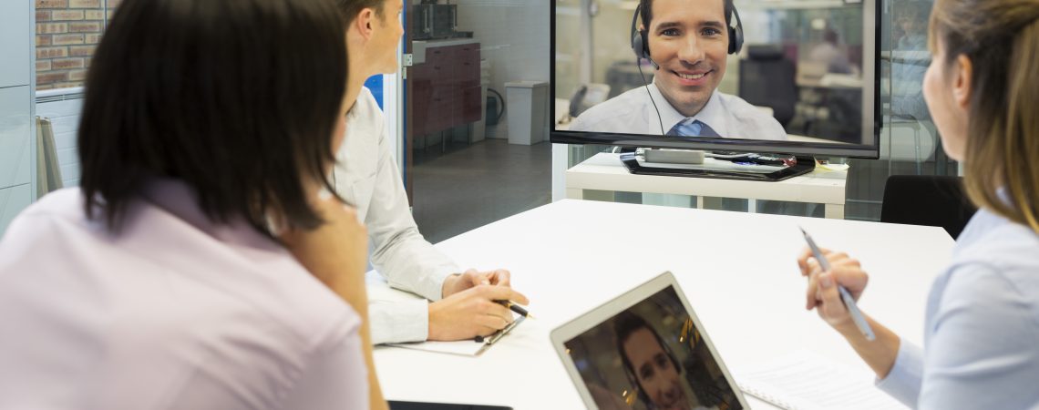 Det sitter tre mennesker rundt et møtebord med laptoper og ser på et digitalt møte på en skjerm
