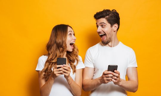 To superhappy unge mennesker med mobil som ser opprømt på hverandre mot oransje bakgrunn