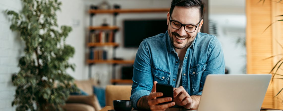 Blid mann ved stuebord med laptop, mobil og kaffekop