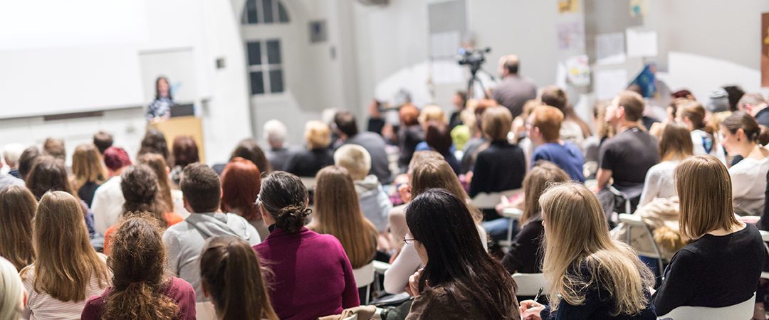 Publikum sett bakfra i en sal hvor konferansieren står foran.