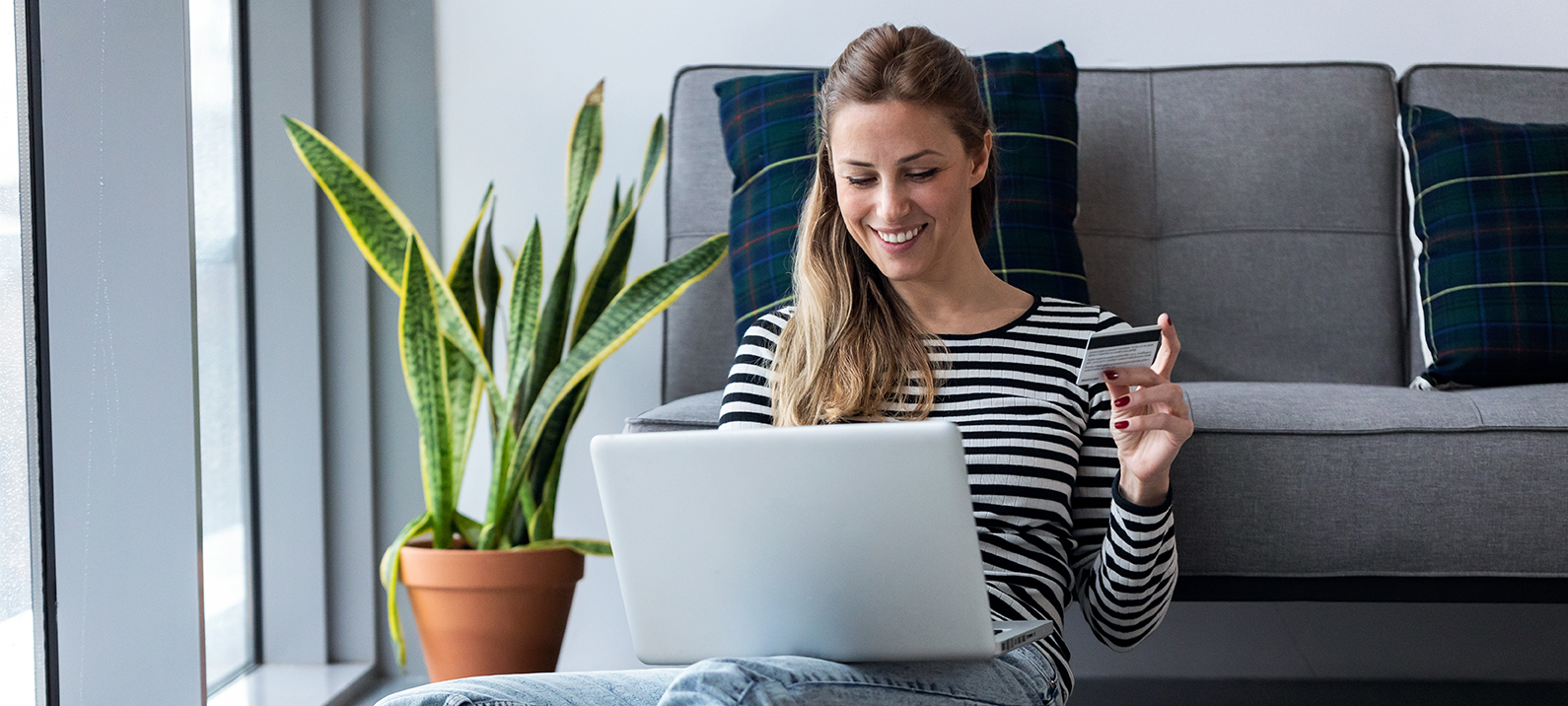 Kvinne som sitter på gulvet foran sofaen med laptop på fanget og betalingskort i hånda.