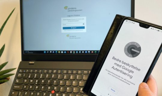 Mobil med forsidebilde av Google Authenticator og laptop med innlogging til Pindena i bakgrunnen