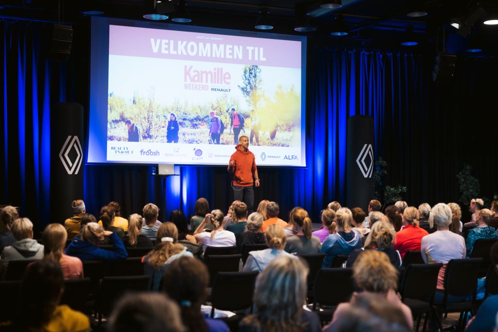 Deltagerne er samlet foran en scene i en sal til Kamilla weekend på Vestlia resort. Konferansier står på scenen foran en velkomstvbilde på storskjerm.