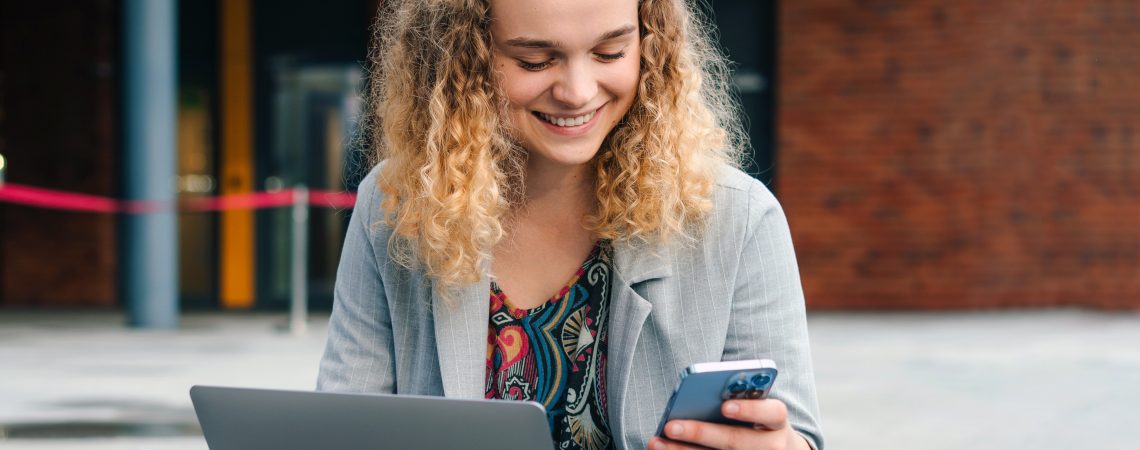 Smilende dame som sitter utenfor et bygg med laptop og mobil