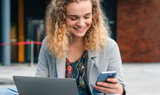 Smilende dame som sitter utenfor et bygg med laptop og mobil