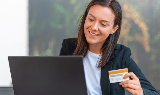 Smilende dame med et bankkort i hånden bak en laptop