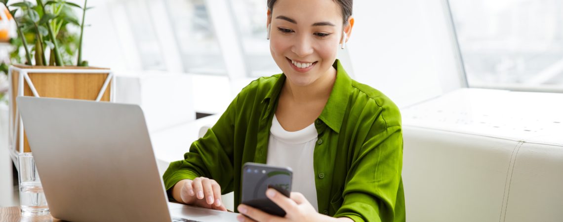 Koordinator som planlegger arrangement i påmeldingssystemet med laptop og mobil ved et skrivebord i en lys stue